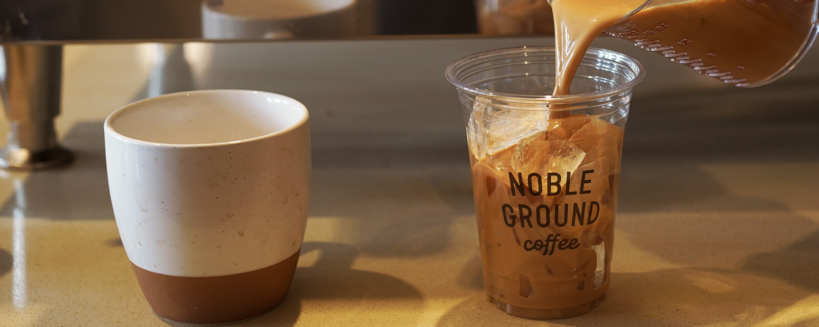 Someone pouring iced coffee in a Noble Grounds coffee cup
